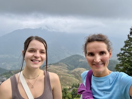 Zwei Frauen lächeln in die Kamera, während sie vor einer beeindruckenden Berglandschaft mit Reisterrassen stehen.