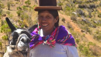 Eine lächelnde Frau mit einem Lama, gekleidet in traditioneller Kleidung, steht in einer grünen, bergigen Landschaft.