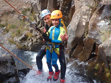 Ein lächelnder Mann und ein Kind stehen in einem Wasserfall, während sie sich an Seilen festhalten und die Natur genießen.