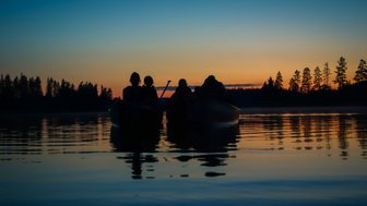 Vier Personen sitzen in Kanus auf einem ruhigen See, während die Sonne hinter den Bäumen untergeht und den Himmel färbt.