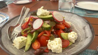 Ein bunter Salat mit Tomaten, Gurken, Paprika und Feta-Käse, serviert auf einem grauen Teller mit Besteck.