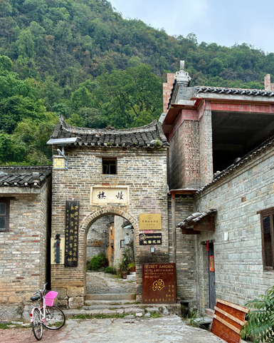 Ein traditionelles Tor führt in eine malerische Gasse, umgeben von alten Steinhäusern und üppiger Natur.