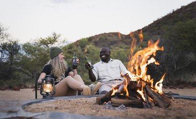 Ein fröhliches Paar sitzt am Lagerfeuer, genießt Getränke und lacht in einer malerischen Umgebung mit sanften Hügeln.
