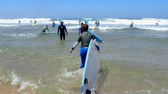 Eine Person mit einem Surfbrett geht ins Wasser, umgeben von anderen Surfern und hohen Wellen unter strahlend blauem Himmel.