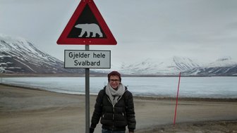 Ein Mann steht vor einem Warnschild, das auf Bären in Svalbard hinweist, mit schneebedeckten Bergen im Hintergrund.