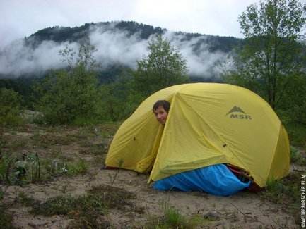 Ein Mann schaut neugierig aus einem gelben Zelt, umgeben von grünen Bäumen und einer nebligen Berglandschaft.