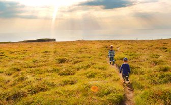 Zwei Kinder wandern auf einem schmalen Pfad durch eine grüne Wiese, während die Sonne über dem Meer aufgeht.