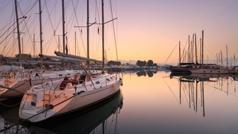 Eine ruhige Marina bei Sonnenaufgang, mit mehreren Segelbooten, die sanft im Wasser schaukeln und ihre Spiegelungen zeigen.