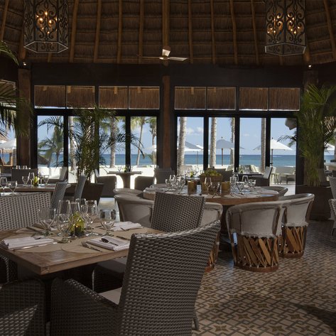 Ein elegantes Restaurant mit runden Holztischen und stilvollen Stühlen, umgeben von Palmen und Meerblick.