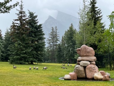 Eine kunstvolle Steinskulptur eines Bären steht im Vordergrund, während Gänse friedlich im Hintergrund grasen.