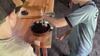 Zwei Kinder stehen an einem Holztisch und mahlen Kaffeebohnen mit einer Handmühle, während eine Holzplatte daneben liegt.