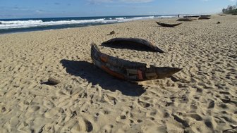 Ein traditionelles Boot liegt im Sand, umgeben von Wellen und einem klaren blauen Himmel, der eine ruhige Atmosphäre schafft.