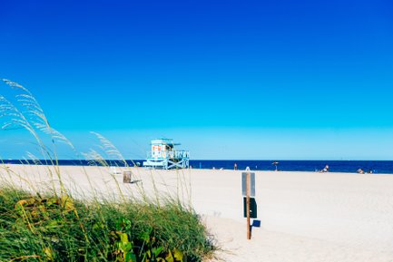 Ein sonniger Strand mit feinem Sand, einem blauen Rettungsturm und Menschen, die sich am Wasser entspannen.