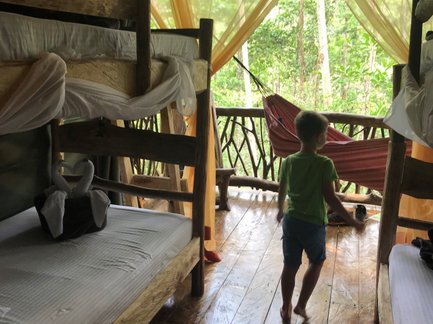 Ein kleiner Junge mit einem grünen T-Shirt läuft barfuß in einem rustikalen Raum mit Holzbetten und Blick auf den Wald.