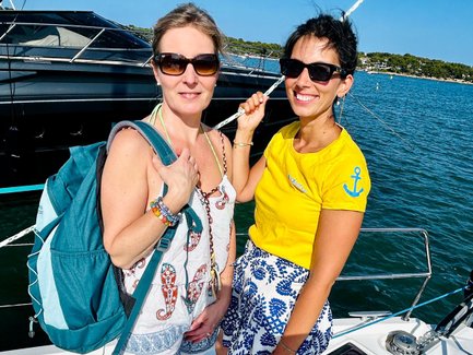 Zwei Frauen stehen auf einem Boot, lächeln und genießen den sonnigen Tag am Wasser, umgeben von einer malerischen Landschaft.