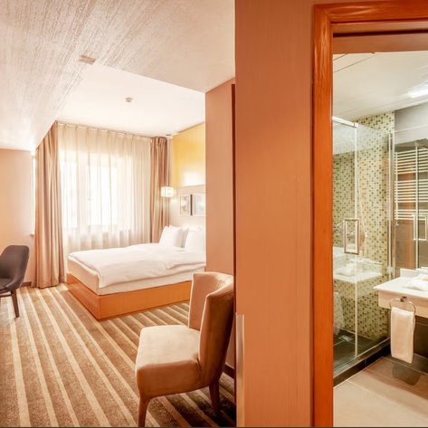 Ein modernes Hotelzimmer mit einem großen Bett, einem Schreibtisch und einem Blick auf das Badezimmer durch eine Glastür.
