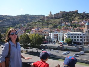 Eine Frau mit Sonnenbrille steht am Rand einer Mauer und blickt auf die Stadt Tiflis mit ihren bunten Häusern und Hügeln.