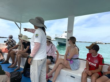 Eine Gruppe von Touristen sitzt auf einem Boot, während eine Führerin auf etwas im Wasser zeigt.