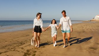 Eine Familie spaziert fröhlich am Strand entlang, während die Sonne am Himmel scheint und das Wasser sanft plätschert.