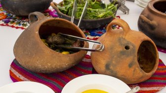 Zwei traditionelle Tontöpfe mit köstlichem Essen stehen auf einem bunten Tisch, umgeben von frischen Salaten und Saucen.