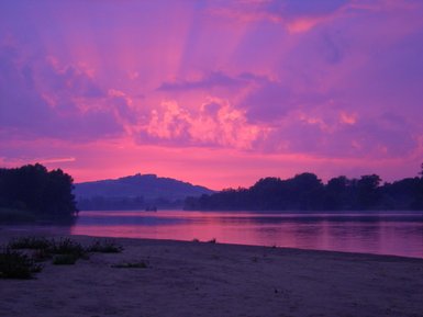 Ein malerischer Sonnenuntergang über einem ruhigen Fluss, umgeben von sanften Hügeln und üppigem Grün im Vordergrund.