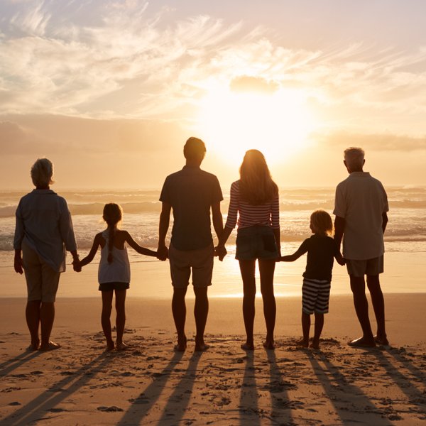 Eine mehrgenerationale Familie steht am Strand und beobachtet den Sonnenuntergang, während die Wellen sanft ans Ufer rollen.