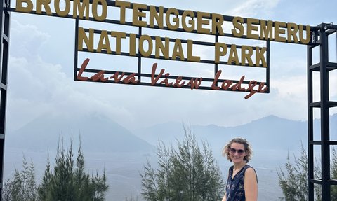 Eine Frau mit lockigem Haar steht lächelnd vor einem Schild des Bromo Tengger Semeru Nationalparks.