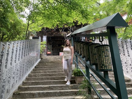 Eine junge Frau steht auf einer steinernen Treppe, umgeben von grünen Bäumen und einem Tempel im Hintergrund.