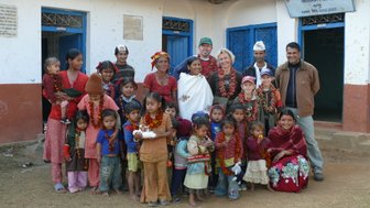 Eine Gruppe von Kindern und Erwachsenen steht vor einem Schulgebäude in Nepal, alle tragen bunte Blumenkränze um den Hals.