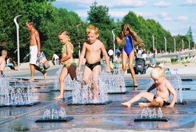 Vier Kinder spielen fröhlich in einem Wasserspielplatz, während Wasserfontänen um sie herum spritzen und die Sonne scheint.