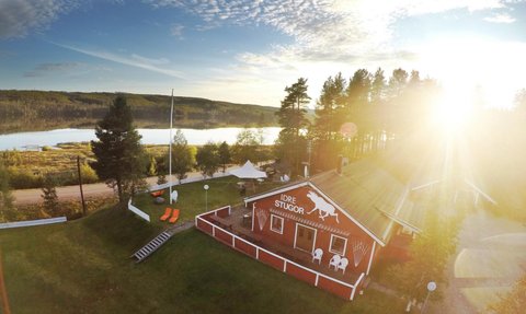 Ein rotes Ferienhaus mit einem weißen Pferdemotiv steht in einer malerischen Landschaft am Wasser, umgeben von Bäumen.