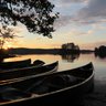 Am Ufer eines ruhigen Sees liegen mehrere Kanus, während die Sonne hinter den Bäumen untergeht und den Himmel erleuchtet.