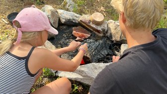 Ein Mädchen in einem gestreiften Shirt grillt über einem Lagerfeuer, während ein Junge daneben zuschaut und sich konzentriert.