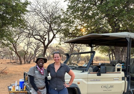 Zwei Personen stehen lächelnd vor einem Geländewagen in der afrikanischen Savanne, umgeben von kahlen Bäumen und sanftem Licht.