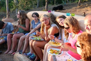 Eine Gruppe von Jugendlichen und Erwachsenen genießt ein gemeinsames Essen im Freien, umgeben von Bäumen und Natur.