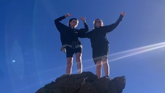 Zwei Personen stehen auf einem großen Felsen und zeigen mit den Händen ein Peace-Zeichen, umgeben von klarem Himmel.