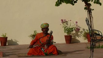 Ein indischer Straßenmusiker sitzt im Freien, trägt ein leuchtendes orangefarbenes Gewand und spielt ein traditionelles Instrument.