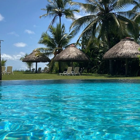Ein glitzernder Pool spiegelt die Sonne wider, umgeben von Palmen und Strohdächern, während eine Person entspannt im Wasser schwimmt.