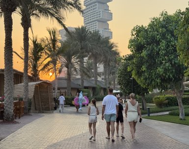 Eine Familie mit zwei Kindern spaziert auf einem gepflasterten Weg, umgeben von Palmen und einem Sonnenuntergang im Hintergrund.