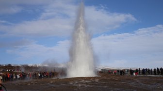 Ein Geysir sprudelt kraftvoll in die Luft, während eine Menschenmenge fasziniert zuschaut und die Wolken am Himmel vorbeiziehen.