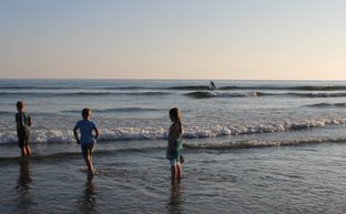 Drei Kinder stehen im flachen Wasser, während sanfte Wellen an den Strand rollen und die Sonne am Horizont scheint.