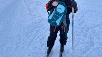 Eine Person steht auf Skiern im Schnee, trägt eine orangefarbene Jacke und hält einen Rucksack in der Hand.