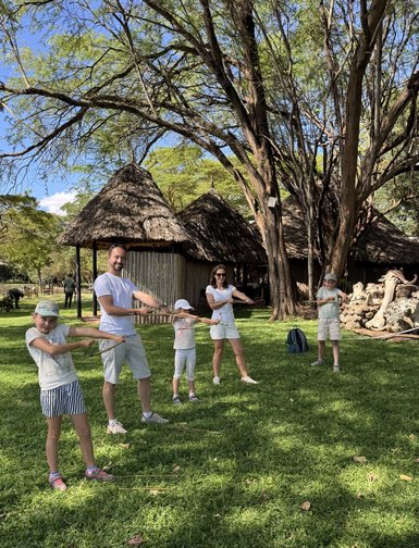Eine Familie steht im Freien und hält Stöcke, umgeben von Bäumen und traditionellen Hütten im Hintergrund.