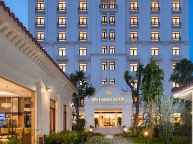 Ein elegantes Hotelgebäude mit dem Namen 'Ninh Binh Hidden Charm' strahlt in der Dämmerung, umgeben von üppigem Grün.