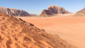 Eine weite Wüstenlandschaft mit sanften Sanddünen und beeindruckenden Felsformationen unter klarem blauen Himmel.
