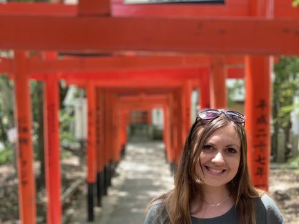 Eine Frau mit langen Haaren steht lächelnd vor einer Reihe von leuchtend roten Torii-Toren in einem ruhigen Park.