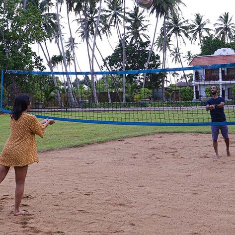 Zwei Personen spielen Volleyball auf einem Sandplatz, umgeben von Palmen und einem Resort im Hintergrund.