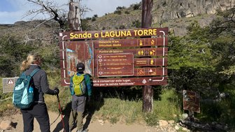 Zwei Wanderer betrachten ein Informationsschild, das den Weg zur Laguna Torre in Patagonien beschreibt, umgeben von Natur.