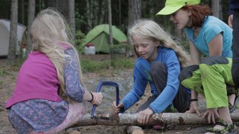 Drei Kinder arbeiten gemeinsam an einem Baumstamm, während sie im Wald campen, um ein Projekt zu starten.