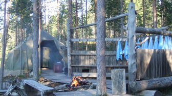 Ein Zeltlager in einem Wald mit einem rauchenden Lagerfeuer und blauen Handtüchern, die an einem Holzgestell hängen.